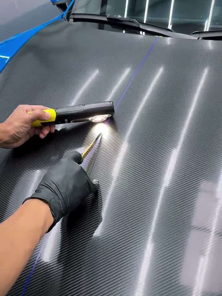 Gloved hand guiding a squeegee across carbon fiber under a work lamp