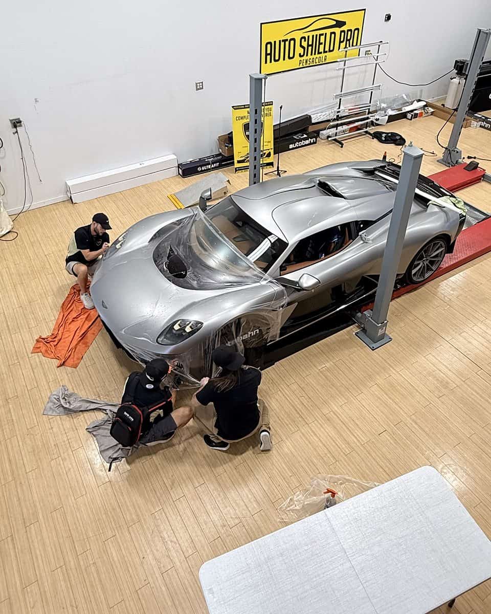 From above: the silver Aston Martin Valhalla on a lift under the Auto Shield Pro Pensacola sign, three people crouched at its front corners