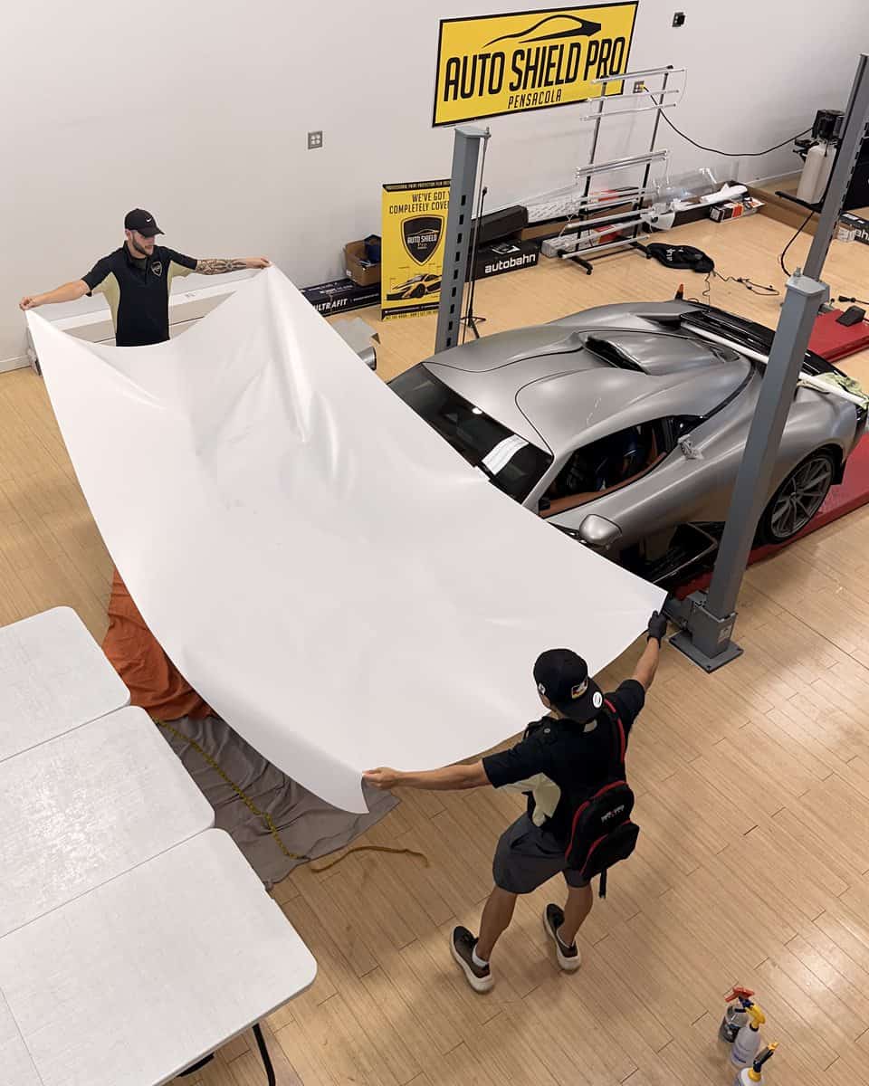 Overhead view of the shop floor: the silver Valhalla on the lift with a white sheet spread open next to it