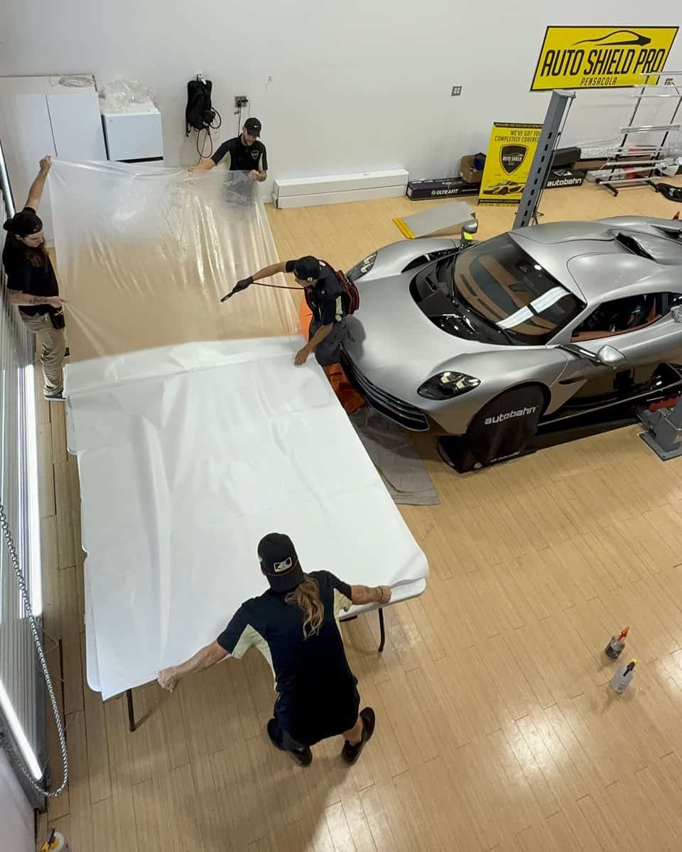 From above: four people at a row of white-covered tables, two holding a clear sheet up while one aims a sprayer at it